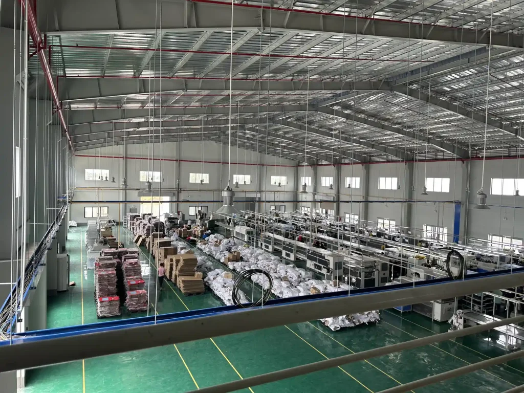 Xưởng sản xuất sàn epoxy xanh với dãy bàn chuyền dài và đèn chiếu sáng treo dưới mái kèo thép